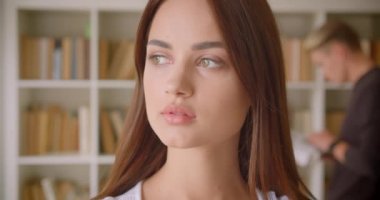 Kütüphanede neşeyle gülümseyen kameraya bakan genç, beyaz beyaz kız öğrencinin arka planda kitap okuyan genç bir erkekle yakın çekim portresi