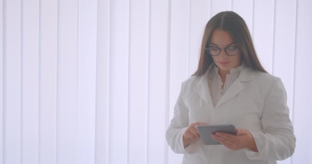 Retrato de close-up de jovem médica branca em óculos usando o comprimido em pé dentro de casa no apartamento branco — Vídeo de Stock