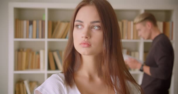 Nahaufnahme Porträt einer jungen hübschen kaukasischen Frau, die in der Bibliothek in die Kamera blickt, während ein junger Mann im Hintergrund ein Buch liest — Stockvideo