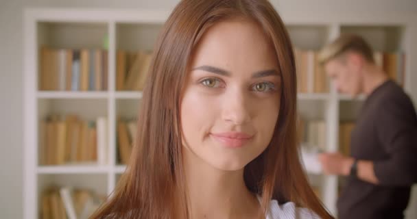 Retrato de cerca de una joven estudiante caucásica mirando a la cámara sonriendo alegremente en la biblioteca con un joven macho leyendo un libro en el fondo — Vídeo de stock