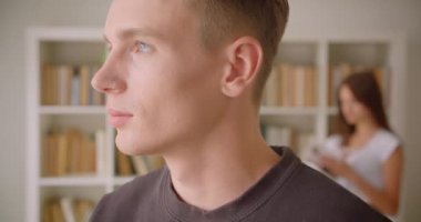 Kütüphanede duran kameraya bakan genç beyaz erkek genç bir kadının arka planda kitap okuduğu yakın çekim portresi