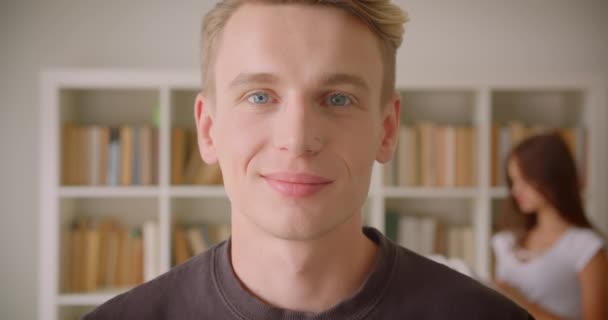 Gros plan portrait de jeune homme caucasien regardant la caméra souriant joyeusement debout dans la bibliothèque avec une jeune femme lisant un livre en arrière-plan — Video