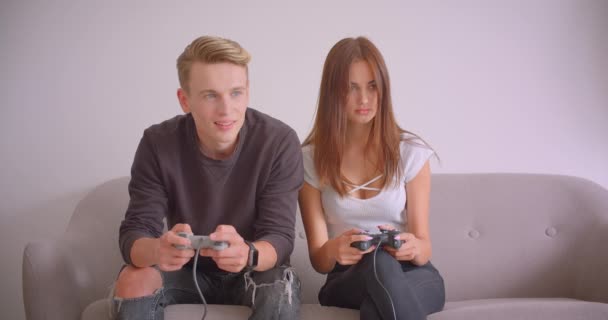 Retrato de close-up de jovem casal bonito caucasiano jogando videogames juntos sentados no sofá dentro de casa no apartamento. Menina conquistando seu namorado — Vídeo de Stock