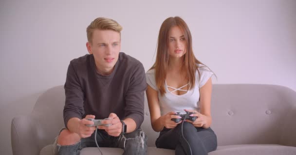 Primer plano retrato de joven linda pareja de jugadores caucásicos jugando videojuegos juntos sentados en el sofá en el interior del apartamento. Chica ganándose a su novio — Vídeo de stock