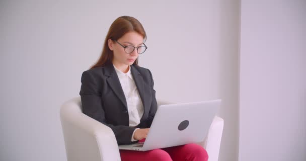 Retrato de cerca de la joven mujer de negocios caucásica en gafas usando el portátil mirando a la cámara sonriendo felizmente sentada en el sillón en el apartamento blanco — Vídeos de Stock