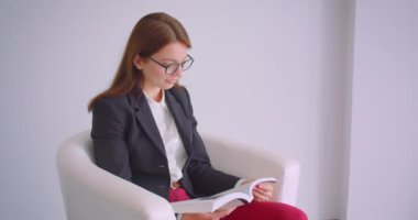 Gözlüklü genç beyaz iş kadınının koltukta mutlu bir şekilde otururken gülümseyen kameraya bakan bir kitap okuduğu yakın çekim portresi