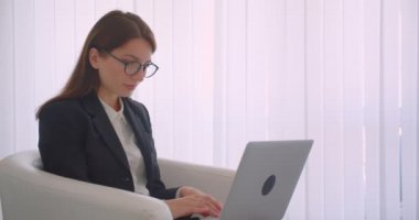 Koltukta mutlu bir şekilde oturan kameraya bakan dizüstü bilgisayar kullanan gözlüklü genç beyaz iş kadınının yakın çekim yan görünümü