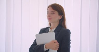 Beyaz ofiste kapalı duran kameraya bakarak dizüstü bilgisayarı tutan genç beyaz iş kadınının yakın çekim portresi