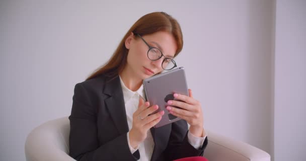 Primer plano retrato de joven mujer de negocios caucásica en gafas de texto en la tableta sentado en el sillón en el apartamento blanco — Vídeos de Stock