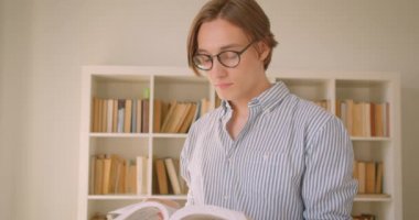 Genç başarılı beyaz erkek öğrencinin gözlüklü yakın çekim portresi kütüphanede kapalı mekanda kameraya bakan bir kitap okuyor