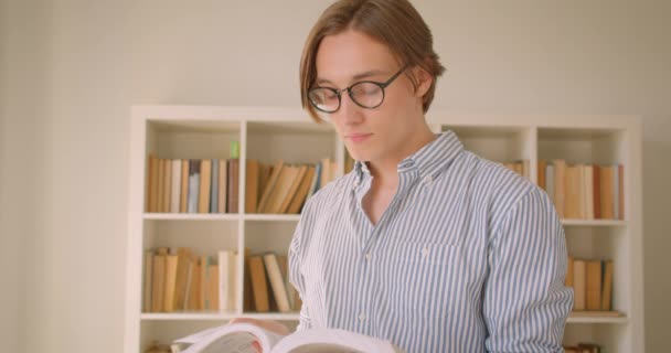 Κοντινό πλάνο του νεαρού επιτυχημένο Καυκάσιο αρσενικό μαθητή σε γυαλιά διαβάζοντας ένα βιβλίο κοιτάζοντας την κάμερα στη βιβλιοθήκη σε εσωτερικούς χώρους — Αρχείο Βίντεο