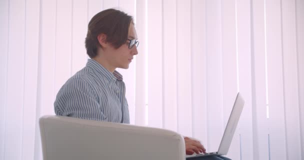 Крупним планом портрет молодого успішного кавказького бізнесмена, який працює на ноутбуці, дивлячись на камеру, що сидить у кріслі в приміщенні — стокове відео
