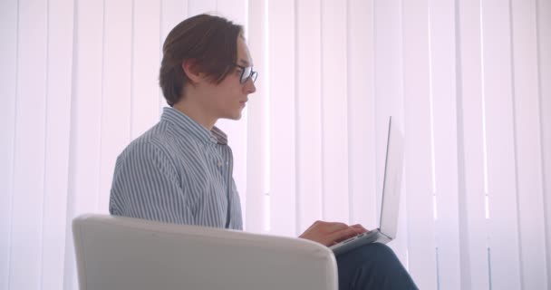 Gros plan côté vue portrait de jeune beau caucasien homme d'affaires travaillant sur l'ordinateur portable regardant caméra assis dans le fauteuil à l'intérieur — Video