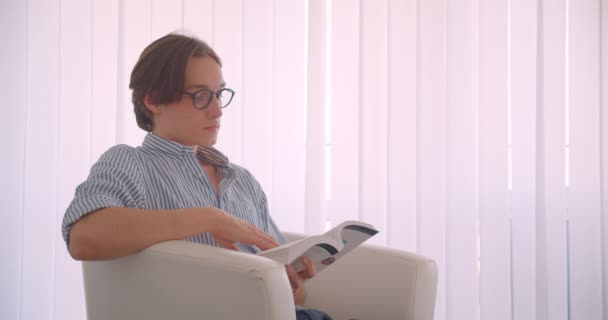 Vista lateral close-up retrato de jovem empresário caucasiano bem sucedido lendo um livro sentado na poltrona dentro de casa — Vídeo de Stock