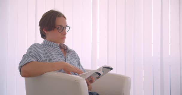 Close-up zijaanzicht portret van jonge succesvolle Kaukasische zakenman het lezen van een boek zittend in de fauteuil binnenshuis — Stockvideo