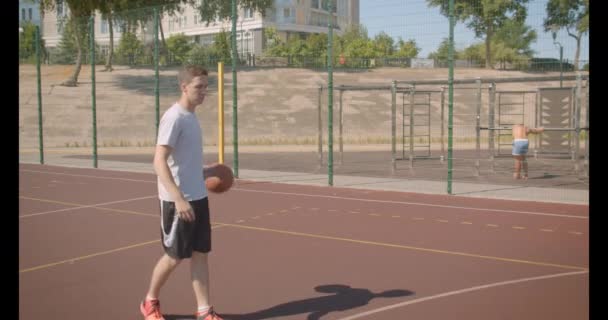 Close-up portret van jonge Kaukasische mannelijke basketbalspeler gooien een bal in de hoepel op het veld buiten — Stockvideo