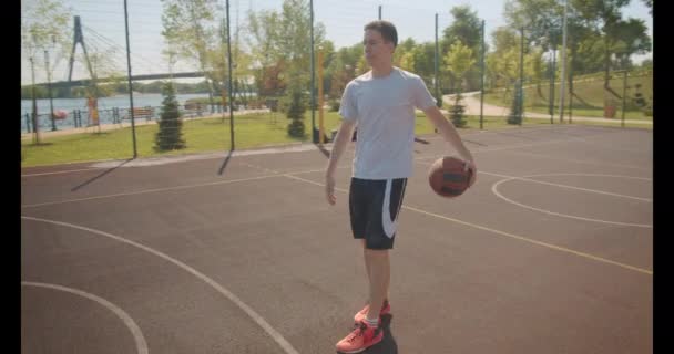 Close-up portret van jonge Kaukasische mannelijke basketbalspeler gooien een bal in de hoepel op de baan met stedelijke gebouwen op de achtergrond — Stockvideo