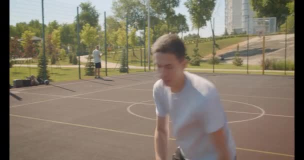 Retrato de primer plano del joven jugador de baloncesto caucásico lanzando una pelota en el aro en la cancha al aire libre con edificios en el fondo — Vídeos de Stock