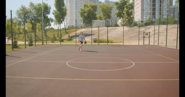 Retrato de primer plano del joven guapo jugador de baloncesto caucásico lanzando una pelota en el aro en la cancha con edificios de la ciudad en el fondo — Vídeos de Stock