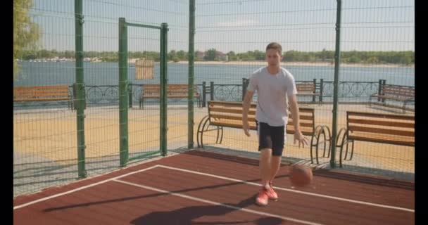 Retrato de primer plano del joven jugador de baloncesto caucásico lanzando una pelota en el aro en la cancha al aire libre junto al río — Vídeos de Stock