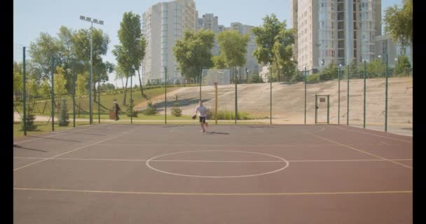 Closeup portrait of young caucasian male basketball player throwing a ball in the hoop on the court with city buildings on the background — Stock Video