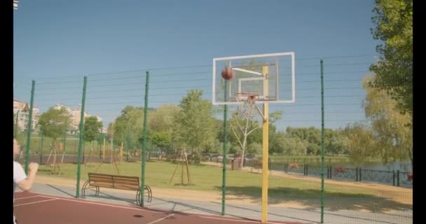 Portrait rapproché d'un jeune joueur de basket-ball caucasien lançant une balle dans le cerceau sur le court à l'extérieur avec des bâtiments urbains en arrière-plan — Video