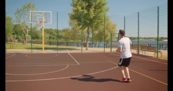 Крупный план портрета молодого привлекательного кавказского баскетболиста, бросающего мяч в кольцо на корте с городскими зданиями на заднем плане — стоковое видео