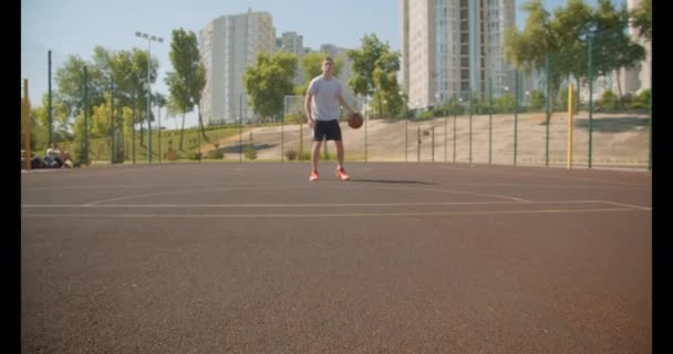 Retrato de primer plano del joven jugador de baloncesto caucásico activo lanzando una pelota en el aro en la cancha con edificios urbanos en el fondo — Vídeo de stock