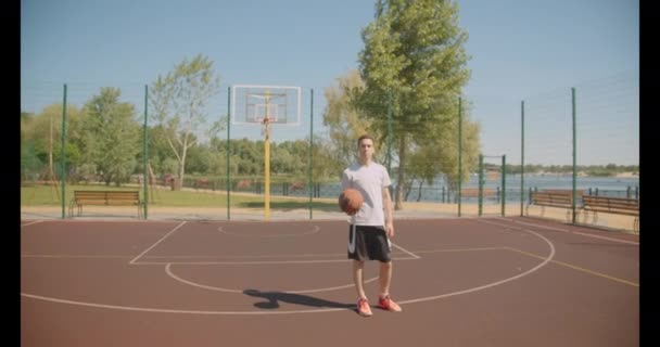 Крупный план портрета молодого кавказского баскетболиста, вращающего мяч на пальце, уверенно смотрящего в камеру на улице с мостом на заднем плане — стоковое видео