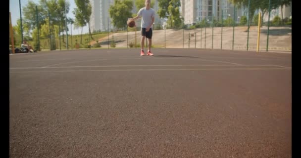 Nahaufnahme Porträt eines jungen aktiven kaukasischen männlichen Basketballspielers, der auf dem Platz einen Ball in den Korb wirft, mit Gebäuden im Hintergrund — Stockvideo