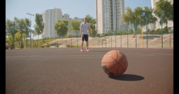 Closeup portrét mladého aktivního kavkazského basketbalového hráče, který hází míč na nádvoří s městskými budovami na pozadí — Stock video