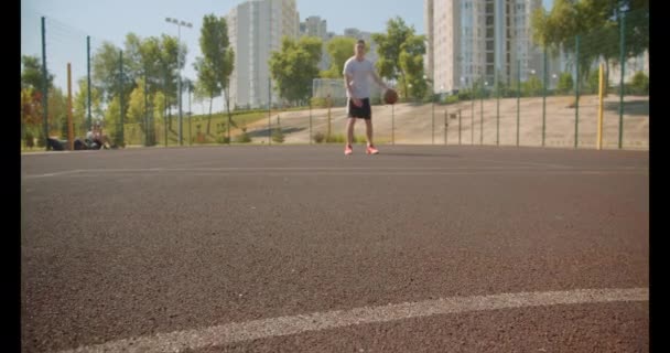 Genç aktif kafkas erkek basketbolcu arka planda kentsel binalar ile sahada pota topu atma Closeup portre — Stok video