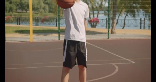 Retrato de primer plano del joven guapo jugador de baloncesto caucásico que sostiene una pelota mirando a la cámara en la cancha al aire libre con puente en el fondo — Vídeo de stock