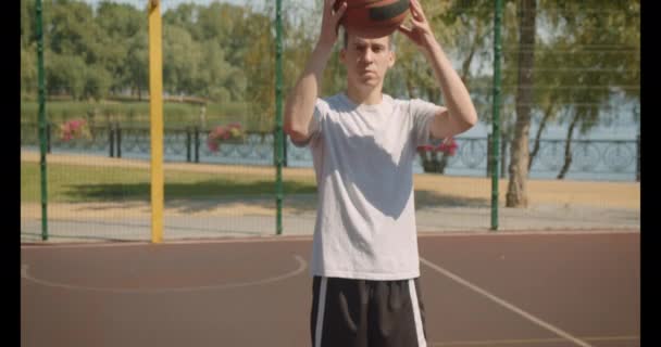 Closeup retrato de jovem branco bonito jogador de basquete masculino segurando uma bola atrás das costas olhando para a câmera na quadra ao ar livre com ponte no fundo — Vídeo de Stock