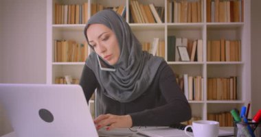 Dizüstü bilgisayar kullanarak başörtüsü genç çekici Müslüman iş kadını Closeup portre ve kapalı kütüphanede notlar alarak telefon görüşmesi olan