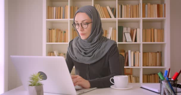 Primer plano retrato de la joven atractiva mujer de negocios musulmana en gafas y hijab utilizando el ordenador portátil y beber café en la biblioteca en el interior — Vídeos de Stock