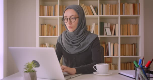 Retrato de cerca de una joven atractiva mujer de negocios musulmana en hijab usando una computadora portátil sentada con satisfacción bebiendo café en la biblioteca dentro — Vídeos de Stock