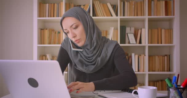 Nahaufnahme Porträt einer jungen attraktiven muslimischen Geschäftsfrau im Hijab, die mit Laptop telefoniert und sich in der Bibliothek Notizen macht — Stockvideo