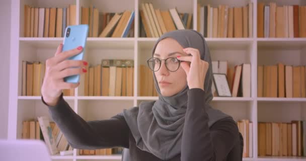 Zbliżenie Portret młodej atrakcyjnej muzułmańskiej bizneswoman w hidżab biorąc selfie na telefon siedzi przed laptopem w bibliotece w pomieszczeniu — Wideo stockowe