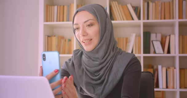 Крупный план портрета молодой привлекательной мусульманки в хиджабе с видеозвонком по телефону, весело разговаривающей сидя перед ноутбуком в библиотеке — стоковое видео