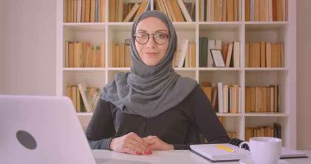 Türbanlı genç çekici Müslüman iş kadınının kapalı mekanda dizüstü bilgisayar kütüphanesinin önünde mutlu bir şekilde gülümseyen kameraya bakan closeup portresi — Stok video