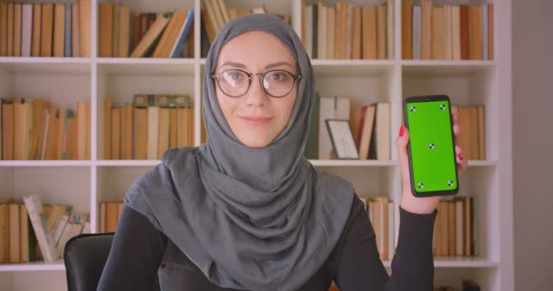 안경을 쓰고 히잡을 착용하고 휴대폰을 사용하여 실내 도서관에서 카메라에 녹색 화면을 보여주는 젊은 매력적인 무슬림 사업가의 클로즈업 초상화 — 비디오