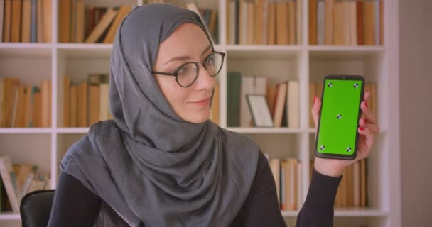 Başörtüsü takan genç Müslüman iş kadınının telefon kullanarak ve kütüphanede kameraya yeşil renk ekran gösteren yakın çekim portresi — Stok video