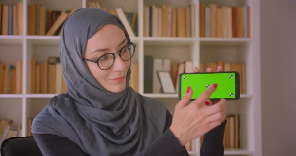 携帯電話を使用してヒジャーブで若いイスラム教徒のビジネスウーマンのクローズアップ肖像画と屋内図書館のカメラに緑のクロマキー画面を示しています — ストック動画