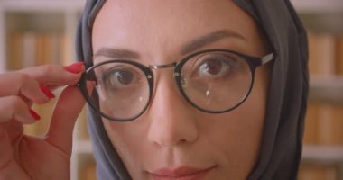 Gözlük ve başörtüsü genç Müslüman kadın yüz closeup portre kapalı kütüphanede neşeyle gülümseyen kameraya bakarak