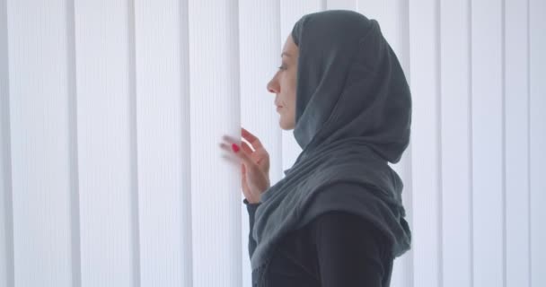 Närbild tillbaka Visa porträtt av unga attraktiva muslimska kvinna i hijab tittar genom fönstret med tyll inomhus i lägenhet — Stockvideo