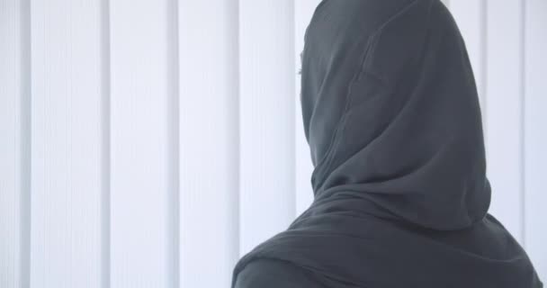 Närbild porträtt av unga attraktiva muslimska kvinnan i hijab tittar genom fönstret med tyll och vänder sig till kameran inomhus i lägenhet — Stockvideo