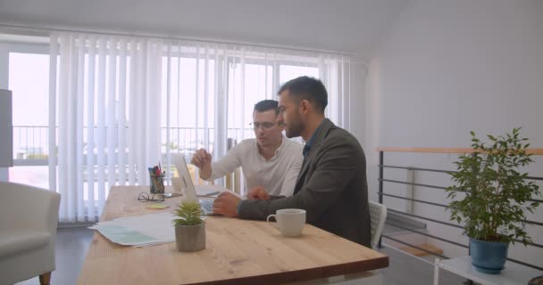 Portret van twee volwassen Kaukasische zakenlui met behulp van de laptop en het hebben van een gesprek in het kantoor binnenshuis — Stockvideo