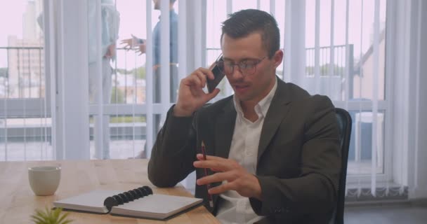 Närbild porträtt av attraktiv affärsman i glasögon som har ett telefonsamtal på kontoret inomhus på arbetsplatsen — Stockvideo