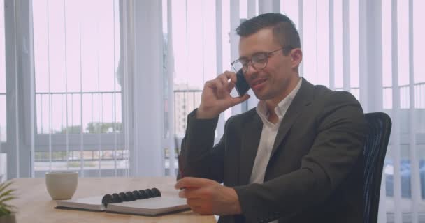 Nahaufnahme Porträt eines attraktiven Geschäftsmannes mit Brille, der im Büro am Arbeitsplatz telefoniert und Notizen macht — Stockvideo